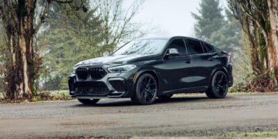 A BMW X6 M parked on a gravel road surrounded by trees and woods.