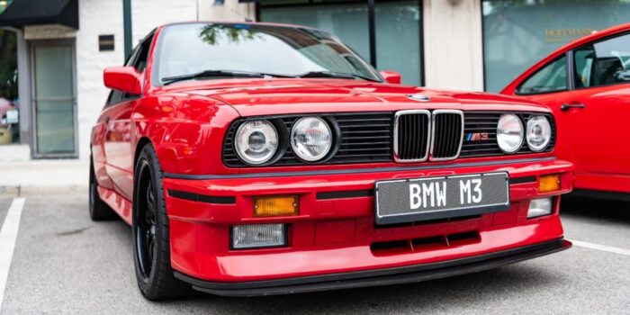 Front view of red BMW E30 M3 parked in an urban setting, showcasing the classic 1980s boxy design with dual round headlights.