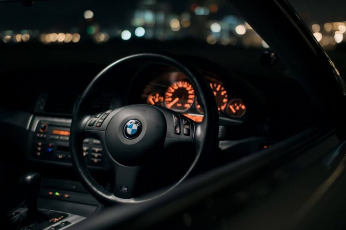 Interior night view of a BMW E30 cockpit with illuminated analog gauges and the iconic BMW steering wheel in focus.