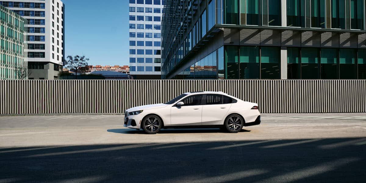 Side profile of a white 2026 BMW 5 series luxury sedan parked in a modern city setting with office buildings in the background