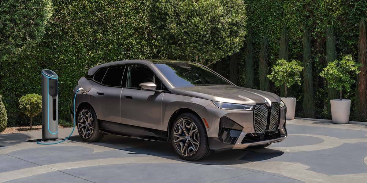 Front three quarter view of a 2026 BMW iX electric SUV parked at a home charging station outdoors in daylight